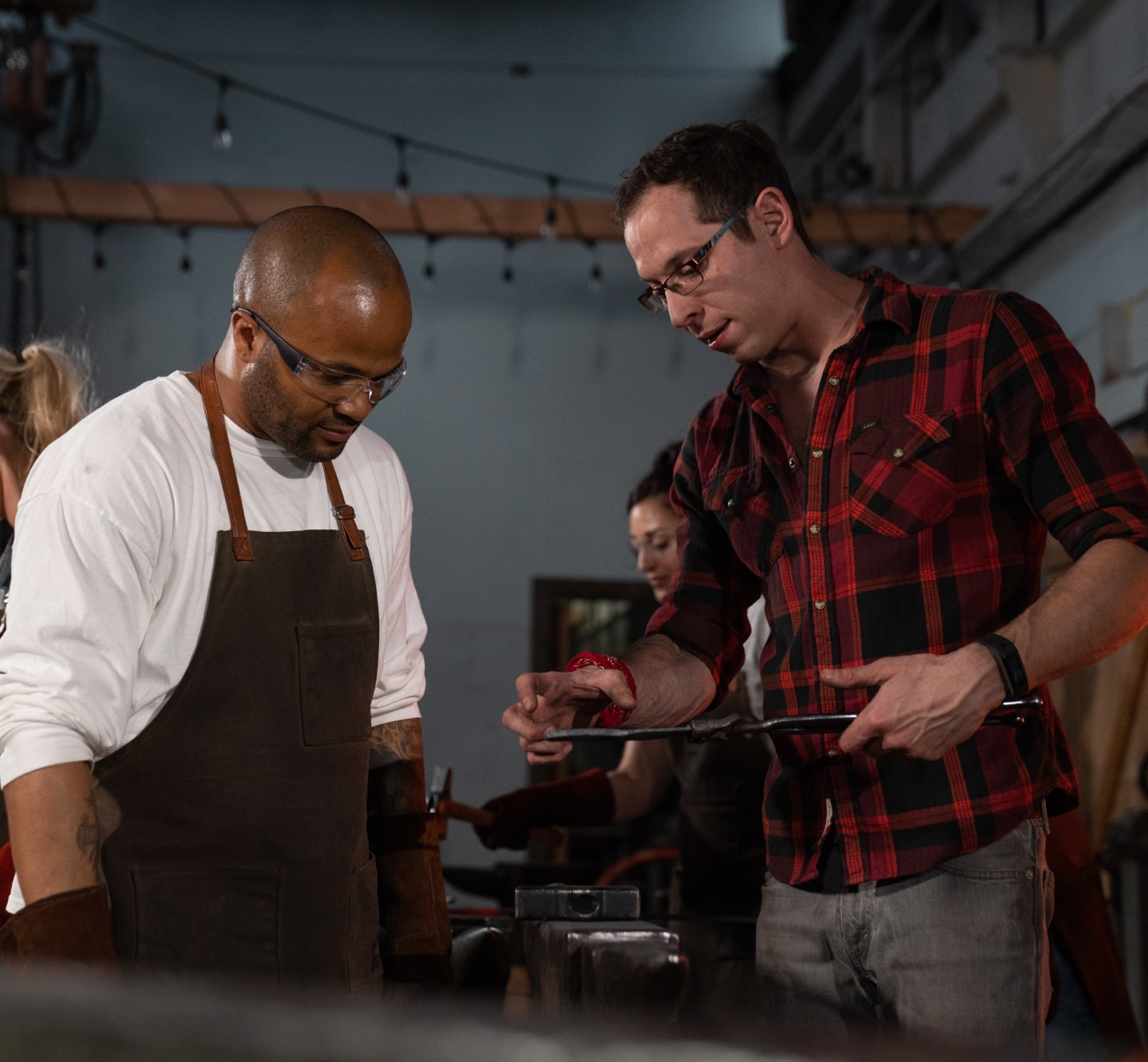 Instructor guiding a student while inspecting a forged metal piece during a blacksmithing workshop.