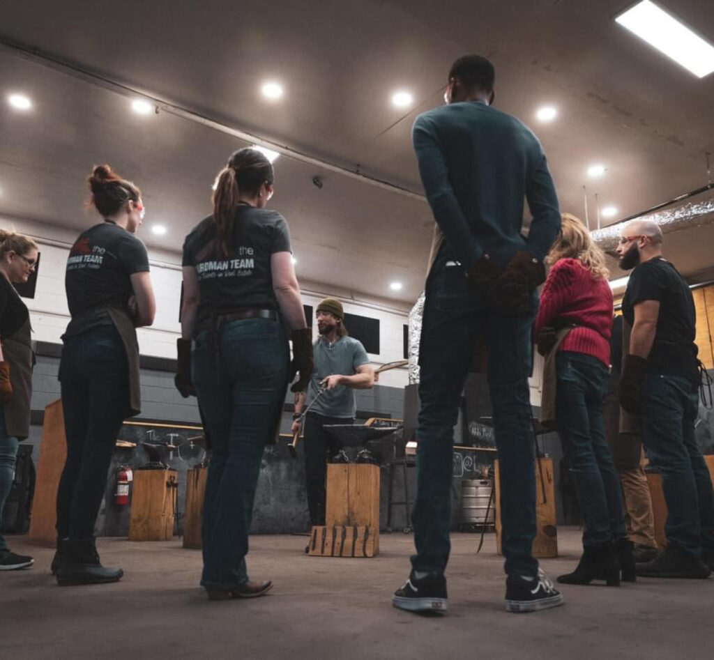 A group of people circles around a blacksmith instructor during a corporate workshop at Lawless Forge