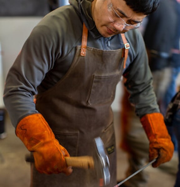 A man in gloves and an apron hammers hot metal during a blacksmithing workshop in Sterling VA