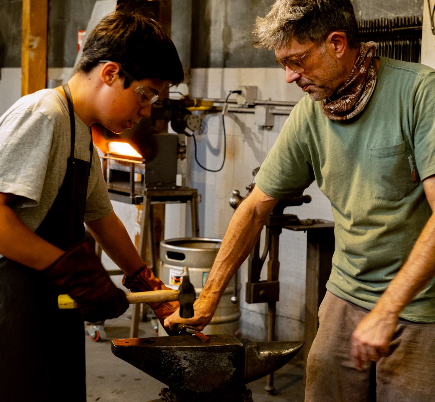 A teenage boy hammers at an anvil while a blacksmith instructor helps him