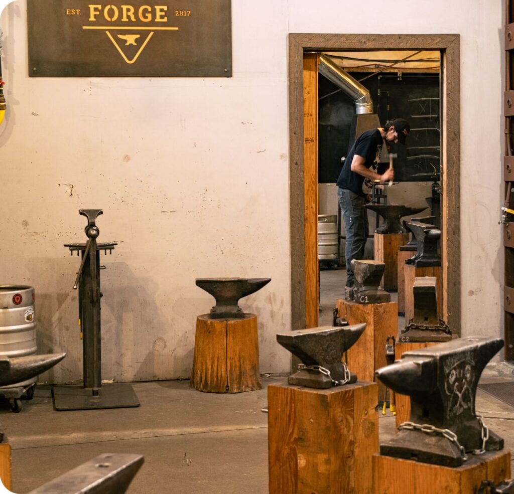 Blacksmith forging hot steel at an anvil inside a blacksmithing workshop surrounded by traditional blacksmithing tools.